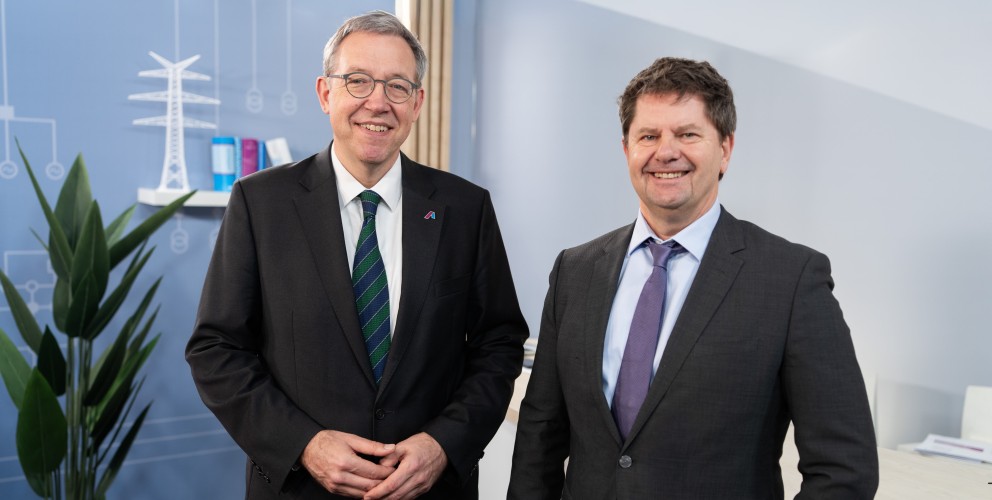 Zwei Männer in dunklen Anzügen stehen nebeneinander und lächeln in die Kamera. Im Hintergrund eine hellblaue Wand mit einer stilisierten Grafik eines Strommasten und einer Windkraftanlage. Bildnachweis: Amprion GmbH / Frank Peterschröder.
