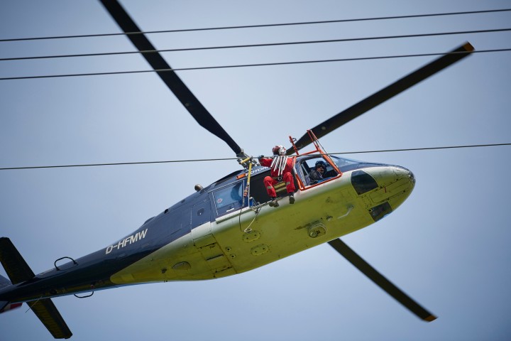 Ein blau/weißer Hubschrauber mit Einziehfahrwerk. In der Tür ein Mensch, er arbeitet an einer Stromleitung.