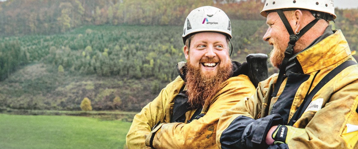 Das Bild im Querformat zeigt zwei Freileitungsmonteure auf einem Hochspannungsmasten. Die beiden Monteure tragen gelbe Kleidung und einen weißen Helm mit dem Amprion-Logo.