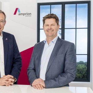 Dr. Christoph Müller und Peter Rüth stehen in Geschäftskleidung in einem Studio. Ihre Hände haben sie jeweils zusammengelegt und locker auf einem weißen Stehtisch abgelegt. Im Hintergrund eine Kulisse mit Amprion Logo und Fenster.