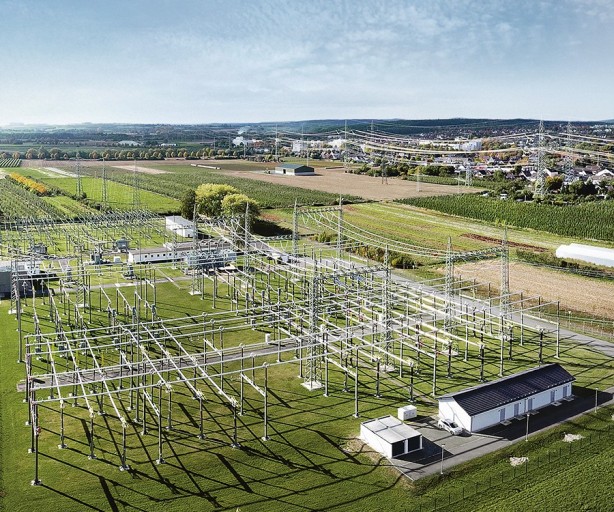 Das Bild zeigt eine luftbildaufnahme eines Umspannwerks in einer ländlichen Umgebung. Im Vordergrund dominiert das Umspannwerk mit seinen charakteristischen, hohen Stahlmasten und zahlreichen Transformatoren. Die Masten sind mit dicken Stromkabeln verbunden, die in einem komplexen Netz angeordnet sind.
Im Hintergrund erstreckt sich eine weitläufige Landschaft mit Feldern und Wiesen. Die Felder sind grün und scheinen kultiviert zu sein, möglicherweise handelt es sich um Ackerland oder Obstplantagen. Am Horizont sind einige Bäume und Gebäude zu erkennen, die auf eine kleine Ortschaft hindeuten.