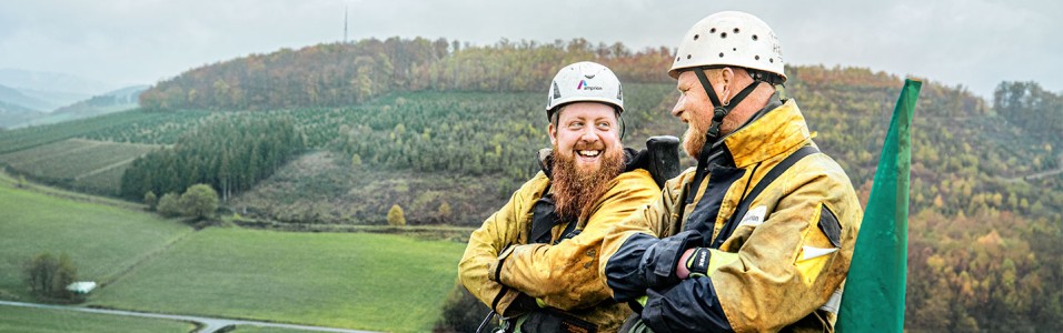 Das Bild zeigt zwei Freileitungsmonteure in gelber Arbeitskleidung mit weißem Helm auf einem Masten in Nahaufnahme.