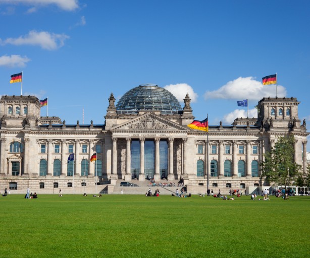 
Dieses Bild zeigt das imposante Reichstagsgebäude in Berlin, Deutschland, aufgenommen an einem sonnigen Tag mit blauem Himmel und einigen Wolken.
Das Gebäude ist im Zentrum des Bildes platziert und dominiert die Ansicht. Es ist ein klassizistisches Gebäude mit einer großen Glaskuppel in der Mitte. Die Fassade ist hell und symmetrisch, mit vielen Fenstern und Säulen, die dem Gebäude eine majestätische Ausstrahlung verleihen. Mehrere deutsche Flaggen wehen an verschiedenen Stellen des Daches und an Masten vor dem Gebäude.
Vor dem Reichstagsgebäude erstreckt sich eine weitläufige, grüne Rasenfläche, der Platz der Republik. Auf dieser Rasenfläche sitzen und liegen einzelne Gruppen von Menschen, die das schöne Wetter genießen. Einige Bäume sind am rechten Bildrand sichtbar und tragen zur natürlichen Umgebung bei.