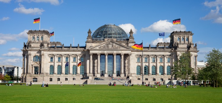 
Dieses Bild zeigt das imposante Reichstagsgebäude in Berlin, Deutschland, aufgenommen an einem sonnigen Tag mit blauem Himmel und einigen Wolken.
Das Gebäude ist im Zentrum des Bildes platziert und dominiert die Ansicht. Es ist ein klassizistisches Gebäude mit einer großen Glaskuppel in der Mitte. Die Fassade ist hell und symmetrisch, mit vielen Fenstern und Säulen, die dem Gebäude eine majestätische Ausstrahlung verleihen. Mehrere deutsche Flaggen wehen an verschiedenen Stellen des Daches und an Masten vor dem Gebäude.
Vor dem Reichstagsgebäude erstreckt sich eine weitläufige, grüne Rasenfläche, der Platz der Republik. Auf dieser Rasenfläche sitzen und liegen einzelne Gruppen von Menschen, die das schöne Wetter genießen. Einige Bäume sind am rechten Bildrand sichtbar und tragen zur natürlichen Umgebung bei.