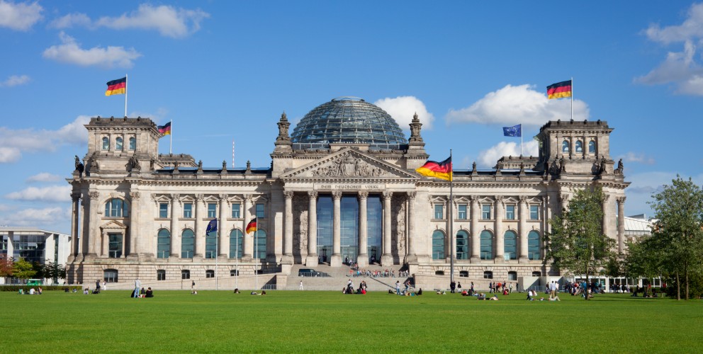 
Dieses Bild zeigt das imposante Reichstagsgebäude in Berlin, Deutschland, aufgenommen an einem sonnigen Tag mit blauem Himmel und einigen Wolken.
Das Gebäude ist im Zentrum des Bildes platziert und dominiert die Ansicht. Es ist ein klassizistisches Gebäude mit einer großen Glaskuppel in der Mitte. Die Fassade ist hell und symmetrisch, mit vielen Fenstern und Säulen, die dem Gebäude eine majestätische Ausstrahlung verleihen. Mehrere deutsche Flaggen wehen an verschiedenen Stellen des Daches und an Masten vor dem Gebäude.
Vor dem Reichstagsgebäude erstreckt sich eine weitläufige, grüne Rasenfläche, der Platz der Republik. Auf dieser Rasenfläche sitzen und liegen einzelne Gruppen von Menschen, die das schöne Wetter genießen. Einige Bäume sind am rechten Bildrand sichtbar und tragen zur natürlichen Umgebung bei.