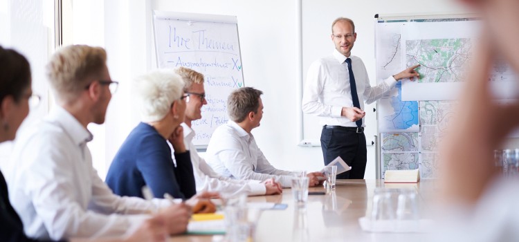 Im Vordergrund ist ein großer, heller Holztisch zu sehen, an dem mehrere Personen sitzen. Die Perspektive ist leicht unscharf und konzentriert sich auf die Mitte des Raumes. Auf dem Tisch stehen Gläser mit Wasser.
Auf der linken Seite des Tisches sitzen drei Personen, deren Gesichter teilweise sichtbar sind. Sie sind aufmerksam dem Redner zugewandt. Die Person im Vordergrund links ist nur von hinten zu sehen.
Rechts in der Mitte des Bildes steht ein Mann mittleren Alters, der die Präsentation hält. Er trägt ein weißes Hemd mit einer dunklen Krawatte und eine Brille. Er gestikuliert mit der rechten Hand auf eine große Tafel oder ein Whiteboard, auf der eine Karte oder ein Plan mit verschiedenen Markierungen und Texten abgebildet ist.