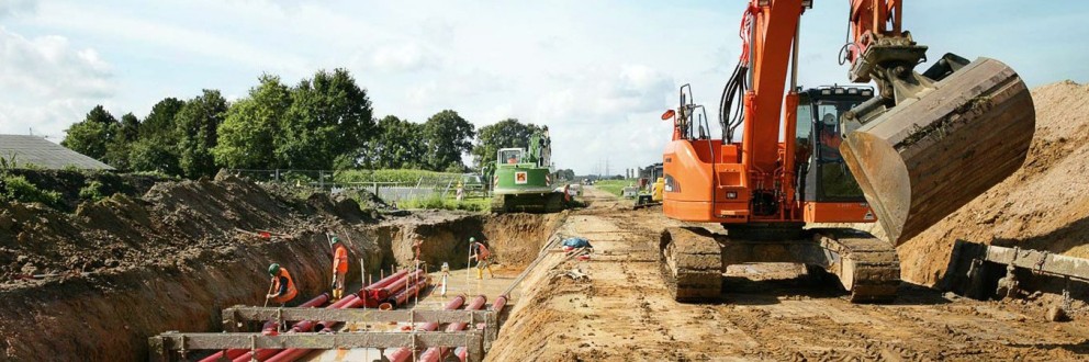 Es folgt eine Bildbeschreibung:
Das Foto im Querformat zeigt eine Erdkabelbaustelle. Links im Bild stehen Arbeiter im Graben, durch den sechs rote Schutzrohre verlaufen. Rechts neben dem kabelgraben steht ein Bagger.
Ende der Bildbeschreibung.
