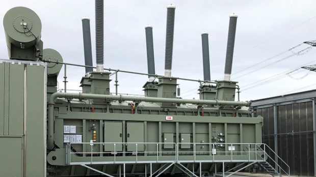 Das Bild zeigt einen großen, industriellen Leistungstransformator auf einem Umspannwerk. Der Transformator ist olivgrün und mit mehreren senkrecht aufragenden, grauen Isolatoren ausgestattet. Diese hohen, zylinderförmigen Isolatoren leiten den elektrischen Strom in das Hochspannungsnetz. An der Seite des Transformators befinden sich diverse Leitungen, Schalter und technische Beschriftungen. Eine Metallplattform mit Treppe ermöglicht den Zugang zu den Wartungseinrichtungen. Im Hintergrund sind weitere elektrische Anlagen sowie Hochspannungsleitungen zu sehen. Der Himmel ist bewölkt.