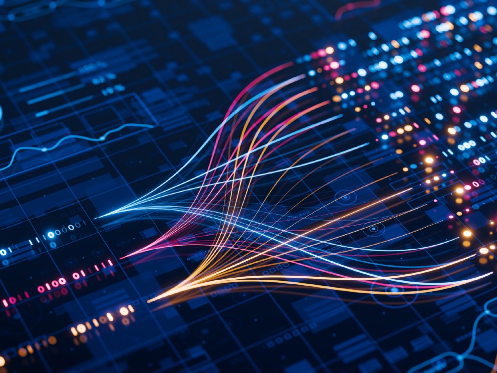 Auf dem Bild ist eine digitale, abstrakte Darstellung zu sehen, die Datenanalyse und Technologie symbolisiert. Der Hintergrund ist in dunklem Blau gehalten und zeigt schemenhaft Graphen, Diagramme und binären Code. Im Zentrum des Bildes erstrecken sich mehrere leuchtende, geschwungene Linien von links nach rechts. Sie beginnen als dünne, hellblaue Kurven, fächern sich auf und enden in einem Strom aus farbigen, leuchtenden Punkten. Die Farben der Kurven und Punkte reichen von Weiß über Blau und Rosa bis hin zu Gelb und Orange.