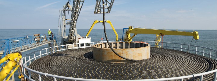 Das Foto im Querformat lässt den Betrachter von oben auf eine große Trommel für Offshore Seekabel blicken. Ringsherum sind Kräne und Gerüste zu erkennen, an den Seiten und dahinter erstreckt sich das Meer.