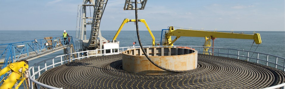 Das Foto im Querformat lässt den Betrachter von oben auf eine große Trommel für Offshore Seekabel blicken. Ringsherum sind Kräne und Gerüste zu erkennen, an den Seiten und dahinter erstreckt sich das Meer.