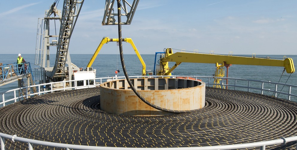 Das Foto im Querformat lässt den Betrachter von oben auf eine große Trommel für Offshore Seekabel blicken. Ringsherum sind Kräne und Gerüste zu erkennen, an den Seiten und dahinter erstreckt sich das Meer.