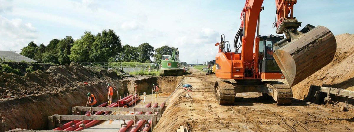 Es folgt eine Bildbeschreibung:
Das Foto im Querformat zeigt eine Erdkabelbaustelle. Links im Bild stehen Arbeiter im Graben, durch den sechs rote Schutzrohre verlaufen. Rechts neben dem kabelgraben steht ein Bagger.
Ende der Bildbeschreibung.
