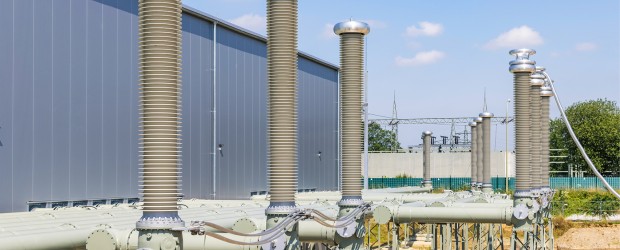 Das Bild zeigt eine Außenansicht einer Umspannanlage oder eines Elektrizitätswerks. Im Vordergrund sind mehrere hohe, säulenartige Isolatoren und dicke Kabelstränge in einem hellgrünen bis grauen Farbton zu sehen. Dahinter erstreckt sich ein großes, graues Gebäude mit einer strukturierten Fassade. Im Hintergrund sind weitere Elemente der Anlage erkennbar, wie Metallgerüste und elektrische Leitungen. Der Himmel ist blau mit einigen weißen Wolken.