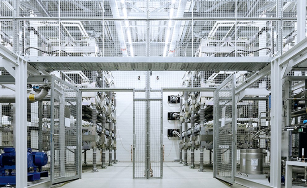 Ein Innenraum einer technischen Anlage mit hohen, metallischen Gestellen, die mit elektrischen Komponenten bestückt sind. Ein engmaschiges Schutzgitter trennt den Gang von den Installationen. Die helle Beleuchtung betont die komplexe Verkabelung und technischen Systeme.
