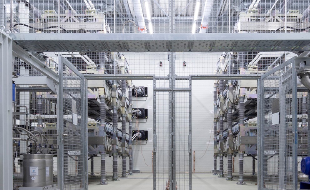 Eine Nahaufnahme des Innenraums einer technischen Anlage mit mehreren parallel angeordneten Schaltschränken und Hochspannungskomponenten. Die Konstruktion ist mit Sicherheitsgittern versehen, während Kabel und Metallstrukturen das Bild dominieren.