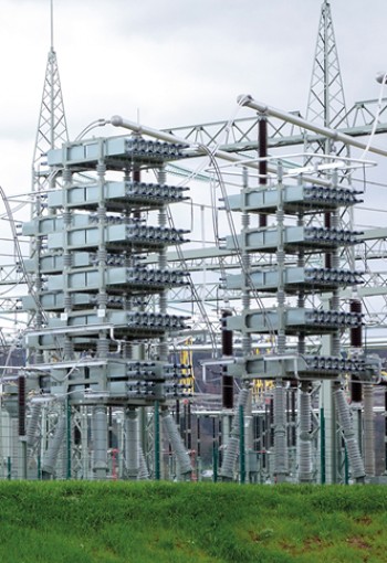 Eine großflächige Freiluftanlage mit mehreren hohen, zylinderförmigen Kompensationsanlagen und metallischen Strukturen. Die Anlage ist von einem Zaun umgeben, mit grüner Wiese im Vordergrund und einem bewölkten Himmel im Hintergrund.
