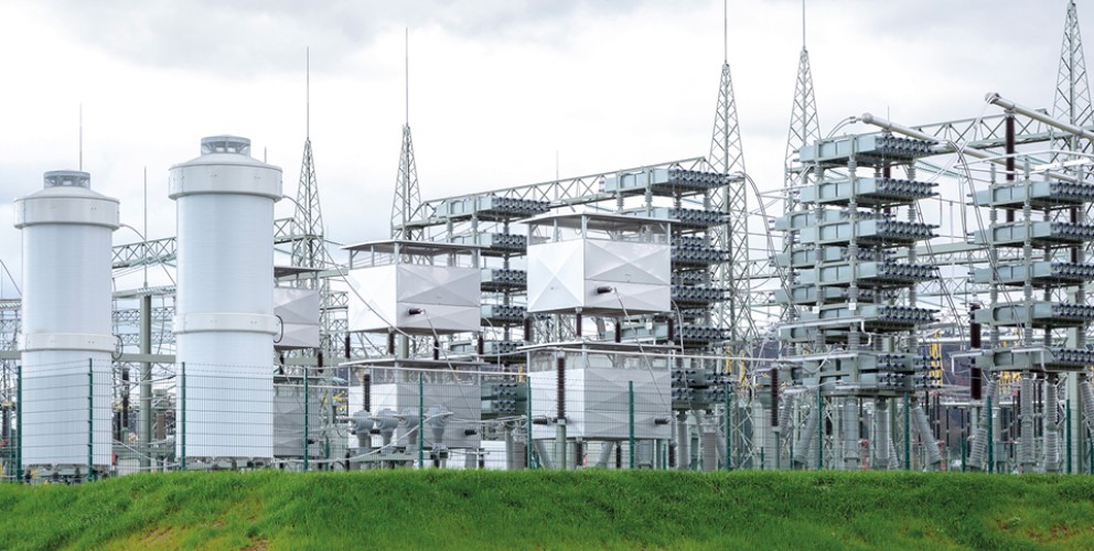 Eine großflächige Freiluftanlage mit mehreren hohen, zylinderförmigen Kompensationsanlagen und metallischen Strukturen. Die Anlage ist von einem Zaun umgeben, mit grüner Wiese im Vordergrund und einem bewölkten Himmel im Hintergrund.
