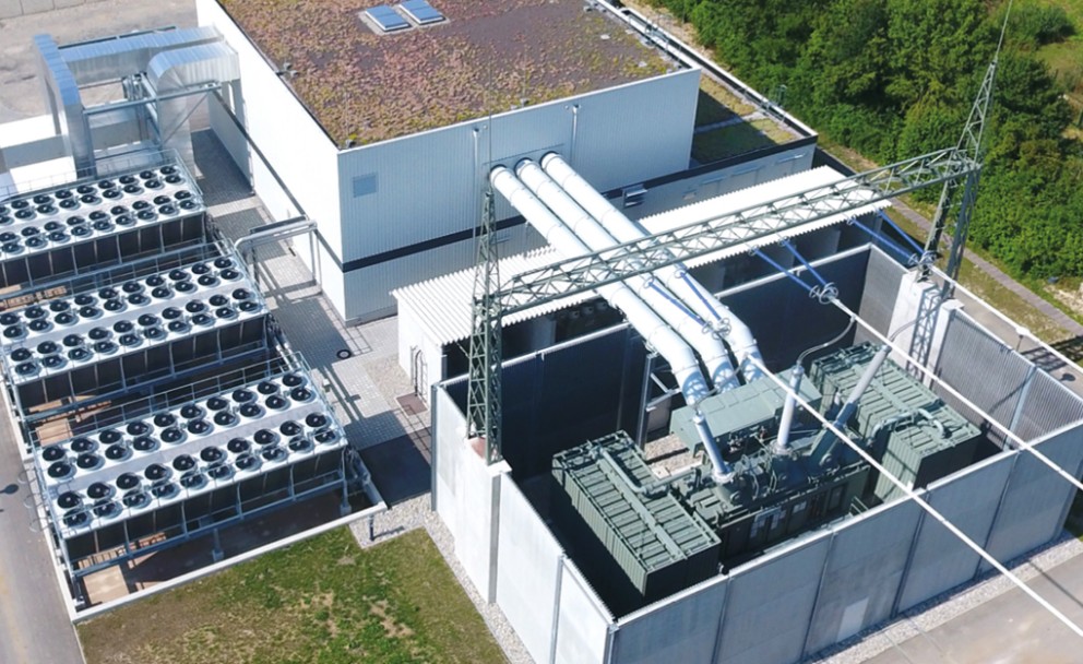 Eine Luftaufnahme einer industriellen Anlage mit einem großen, weißen Gebäude mit begrüntem Dach. Daneben befinden sich mehrere große Kühlanlagen mit Ventilatoren. Im Vordergrund sind massive Transformatoren und Hochspannungsleitungen zu sehen, die in eine Umspannstation führen.