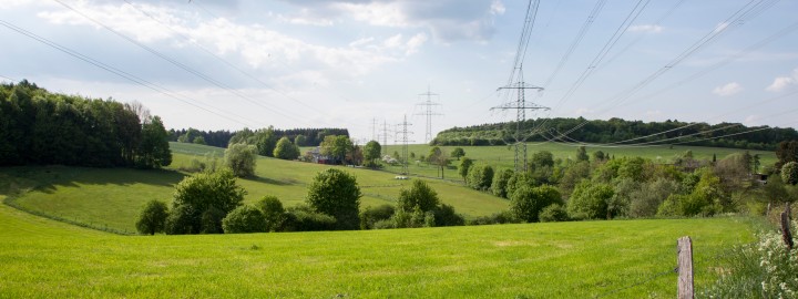 Beginn der Bildbeschreibung:
Auf dem Bild im Querformat ist eine vom Betrachter her in die Ferne laufende Stromtrasse in einer idyllischen hügeligen und grünen Frühlingslandschaft mit Waldsäumen und einzelnen Bäumen sowie Büschen zu erkennen.
Ender der Bildbeschreibung