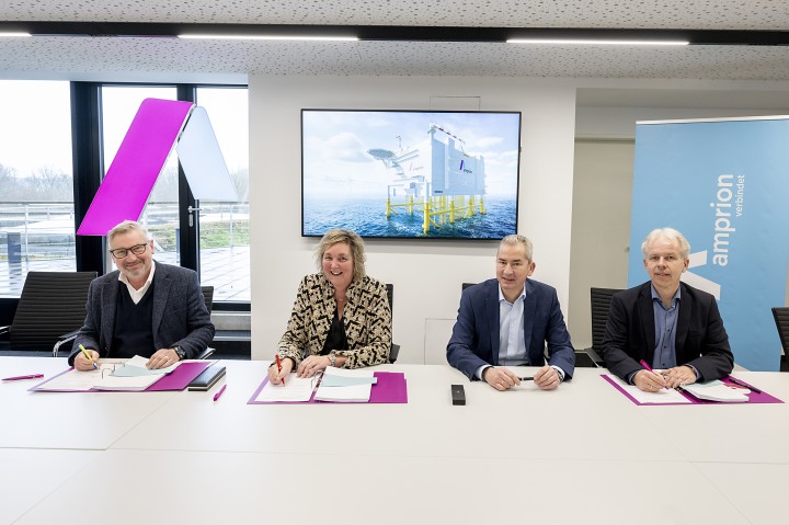Vier Personen sitzen an einem Konferenztisch und unterzeichnen Dokumente, die auf lilafarbenen Mappen liegen. Sie lächeln in die Kamera. Im Hintergrund ist ein Bildschirm zu sehen, auf dem eine Offshore-Konverterplattform mit dem Amprion-Logo über dem Meer dargestellt ist. Links im Raum befindet sich eine moderne Leuchtskulptur in Form eines stilisierten "A" mit lila und weißer Beleuchtung. Rechts steht ein blauer Banner mit dem Amprion-Slogan „verbindet“. Der Konferenzraum hat eine große Fensterfront mit Blick auf eine Terrasse.