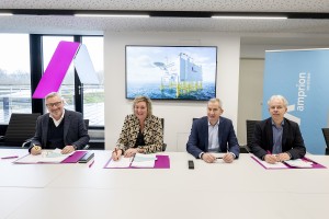 Vier Personen sitzen an einem Konferenztisch und unterzeichnen Dokumente, die auf lilafarbenen Mappen liegen. Sie lächeln in die Kamera. Im Hintergrund ist ein Bildschirm zu sehen, auf dem eine Offshore-Konverterplattform mit dem Amprion-Logo über dem Meer dargestellt ist. Links im Raum befindet sich eine moderne Leuchtskulptur in Form eines stilisierten "A" mit lila und weißer Beleuchtung. Rechts steht ein blauer Banner mit dem Amprion-Slogan „verbindet“. Der Konferenzraum hat eine große Fensterfront mit Blick auf eine Terrasse.