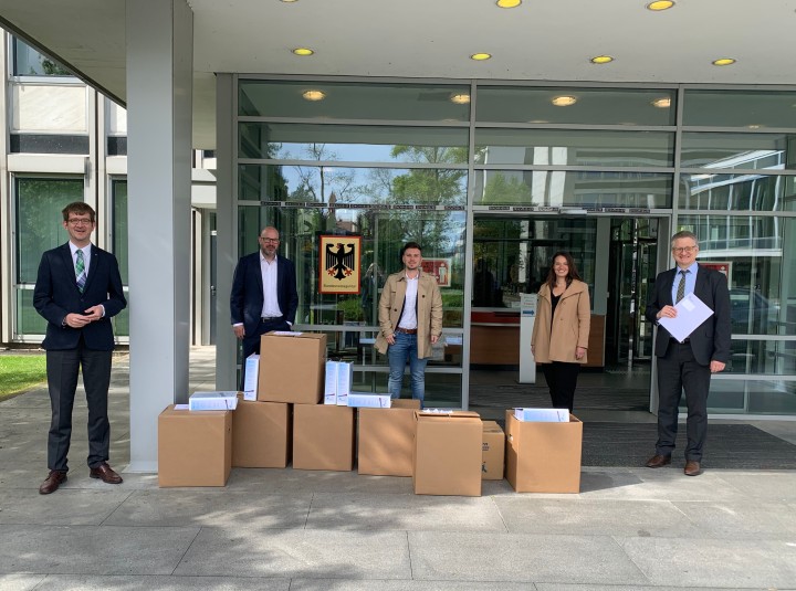 Fünf Personen stehen vor einem modernen Gebäude mit Glasfassade, über dessen Eingang das Schild der Bundesnetzagentur mit dem Bundesadler hängt. Sie sind geschäftlich gekleidet, einige tragen Anzüge, andere Mäntel. Vor ihnen sind mehrere große Kartons aufgestapelt, aus denen Aktenordner und Dokumente herausragen. Zwei der Männer halten ebenfalls Unterlagen in der Hand. Die Gruppe wirkt offiziell und posiert für das Foto, vermutlich im Zusammenhang mit der Übergabe von Dokumenten an die Behörde.