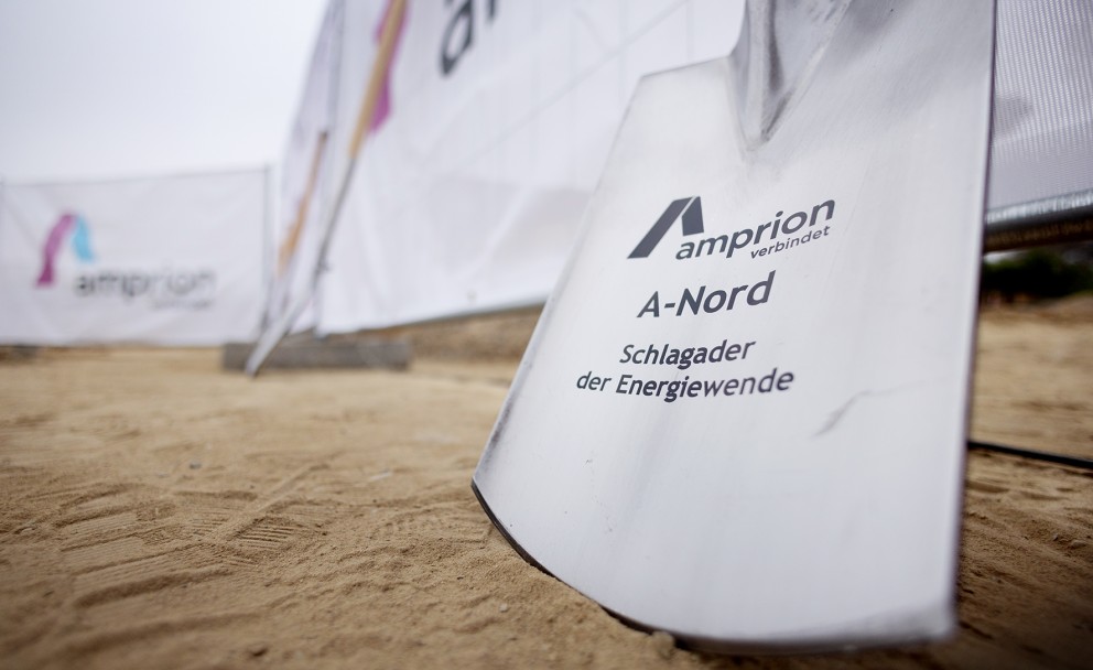 Das Bild zeigt einen silberfarbenen Spaten, der auf einem sandigen oder erdfarbenen Boden steht. Auf dem Spatenblatt ist das Amprion-Logo in Magenta und Schwarz zu sehen, zusammen mit dem Schriftzug "amprion verbindet". Darunter steht in größerer, schwarzer Schrift "A-Nord" und darunter der Slogan "Schlagader der Energiewende" in kleinerer, schwarzer Schrift.