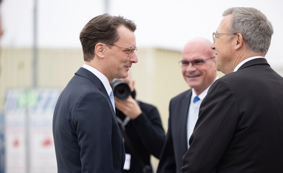 Das Bild zeigt eine Szene mit vier Männern im Freien, die sich zu unterhalten scheinen. Drei der Männer sind im Fokus, während der vierte im Hintergrund teilweise verdeckt ist.
Links im Bild steht ein Mann mit dunklen, leicht gewellten Haaren und Brille. Er trägt einen dunkelblauen Anzug, ein hellblaues Hemd und eine hellblaue Krawatte mit weißen Punkten. Er blickt nach rechts und scheint zuzuhören oder mit den anderen Männern zu sprechen.
In der Mitte steht ein Mann mit einer Glatze und Brille. Er trägt einen dunklen Anzug, ein hellblaues Hemd und eine blaue Krawatte mit weißen Punkten. Er lächelt und blickt ebenfalls nach rechts.
Rechts im Bild steht ein Mann mit kurzen, grauen Haaren und Brille. Er trägt einen dunklen Anzug und ein weißes Hemd. Sein Gesicht ist im Profil zu sehen, er blickt nach links und scheint ebenfalls am Gespräch beteiligt zu sein.