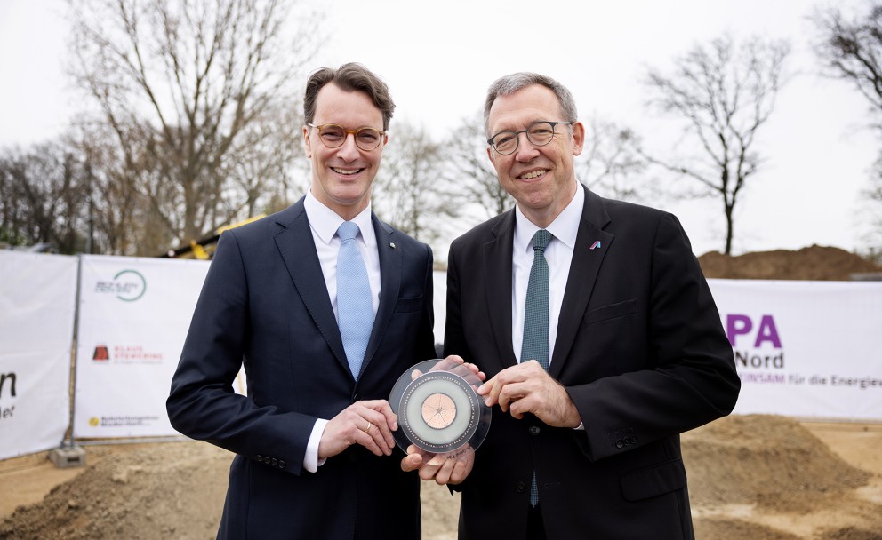 Das Bild zeigt zwei Männer in Anzügen, die nebeneinander stehen und in die Kamera lächeln. Der Mann links hat braune, leicht gewellte Haare und trägt eine Brille, einen dunkelblauen Anzug, ein weißes Hemd und eine hellblaue Krawatte mit weißem Muster. Er hält mit beiden Händen eine transparente, runde Plakette mit einem kupferfarbenen Zentrum und konzentrischen Kreisen.
Der Mann rechts hat graue, kurze Haare und trägt ebenfalls eine Brille, einen schwarzen Anzug, ein weißes Hemd und eine grüne Krawatte mit einem Muster. Er hält die Plakette ebenfalls mit beiden Händen und blickt freundlich in die Kamera.
Im Hintergrund sind unscharf Teile einer Baustelle zu erkennen. Es gibt einen sandigen oder erdfarbenen Boden, einen weißen Bauzaun mit Aufschriften und Logos (teilweise lesbar ist "A-Nord") und einige kahle Bäume. Der Himmel ist grau und bedeckt, was auf einen trüben Tag hindeutet.