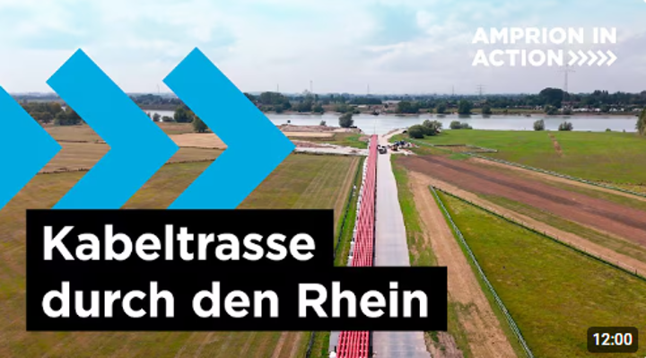 Das Bild zeigt eine Luftaufnahme einer Baustelle für eine Kabeltrasse entlang eines Deichs, der zu einem breiten Fluss führt. 
Visuelle Beschreibung
Vordergrund: Grüne Felder und Ackerland dominieren den unteren und mittleren Bereich. In der Mitte des Bildes verläuft ein breiter, roter Streifen, der die Baustelle der Kabeltrasse markiert und in Richtung des Wassers führt.
Mittelgrund: Der rote Streifen endet an einer Baustellenzufahrt direkt am Ufer eines großen, breiten Flusses (dem Rhein). Auf der gegenüberliegenden Seite des Flusses sind ebenfalls landwirtschaftliche Flächen und im Hintergrund eine Baumreihe sichtbar.
Hintergrund: Der Horizont zeigt eine flache Landschaft mit weiteren Feldern, Bäumen und einigen Strommasten am fernen Ufer. Der Himmel ist hell mit leichter Bewölkung.
Textüberlagerungen:
Links unten in einem schwarzen Kasten steht der weiße Text: "Kabeltrasse durch den Rhein".
Oben rechts in Weiß: "AMPRION IN ACTION >>>>>".
Unten rechts ist in einem schwarzen Kasten eine Zeitangabe: "12:00".
Grafische Elemente: Auf der linken Seite sind große, stilisierte blaue Pfeile platziert, die nach rechts zeigen.
