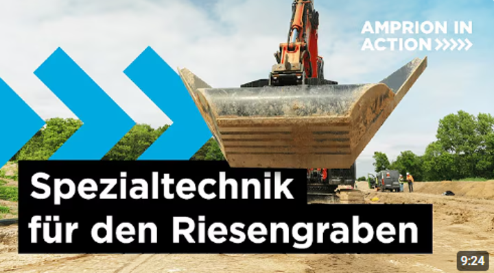 Eine Nahaufnahme zeigt die große, dreckige Baggerschaufel eines orangefarbenen Baggers auf einer Baustelle mit Erdarbeiten. Im oberen rechten Bereich steht der Text 'AMPRION IN ACTION >>>>>>>' und unten im schwarzen Feld der Titel 'Spezialtechnik für den Riesengraben'