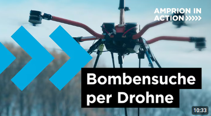 Nahaufnahme einer rot-schwarzen Drohne im Flug. Im unteren Bereich steht der Text Bombensuche per Drohne in weißen Buchstaben auf schwarzem Hintergrund, oben rechts Amprion In Action