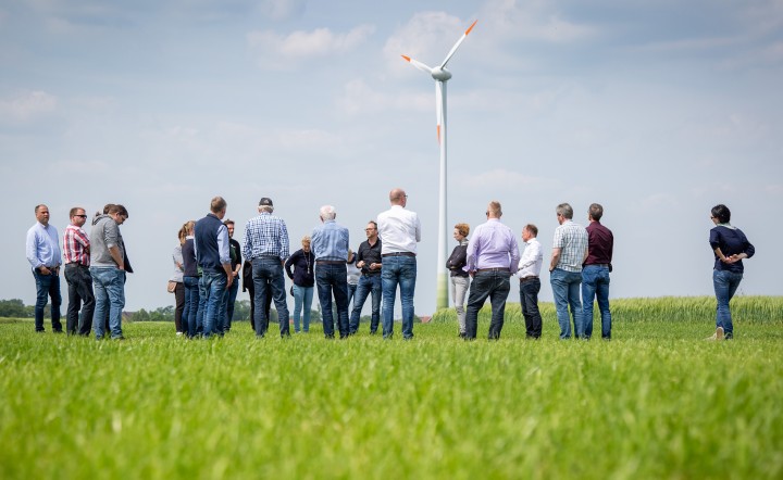 Auf dem Foto steht eine Gruppe Menschen auf einem grünen Rasenstück. Im Hintergrund ein Windrad. Es ist leicht bewölktes Wetter.