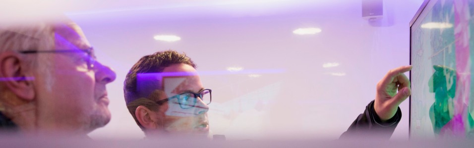 Das leicht lilafarbene Bild zeigt einen Amprion-Mitarbeiter, der einem Bürger an einem Bildschirm die Trassenkorridore im Projekt A-Nord erläutert.