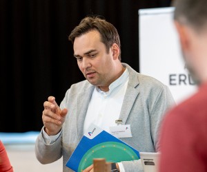 Ein Mann im grauen Sakko und weißen Hemd gestikuliert aufmerksam in einer Gesprächsrunde und hält eine blaue Informationsmappe.