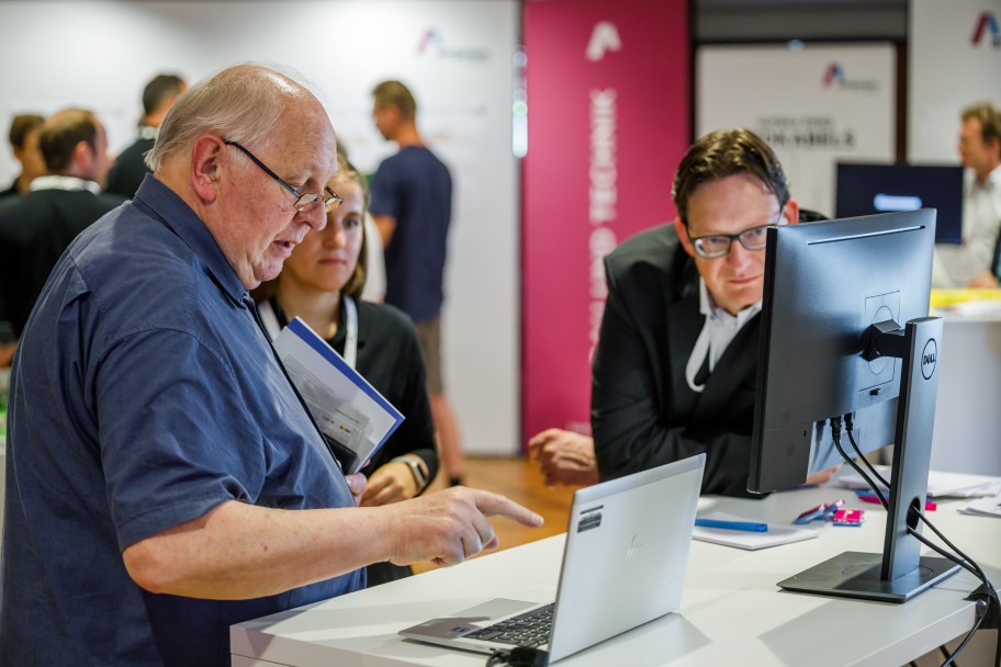 Schnappschuss von einer Infoveranstaltung: Zwei Personen stehen an einem Tisch, auf dem mehrere Computerbildschirme stehen. Eine Person deutet mit dem Zeigefinger auf den aufgeklappten Laptop.