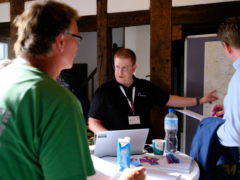 Ein Mann in einem schwarzen Shirt steht vor einer Pinnwand und deutet mit dem Zeigefinger auf die Karte, die aufgehängt ist. Sein Blick geht in die entgegengesetze Richtung. Um ihn herum stehen mehrere Personen, eine davon verschwommen mit Bick zur Karte im Vordergrund.