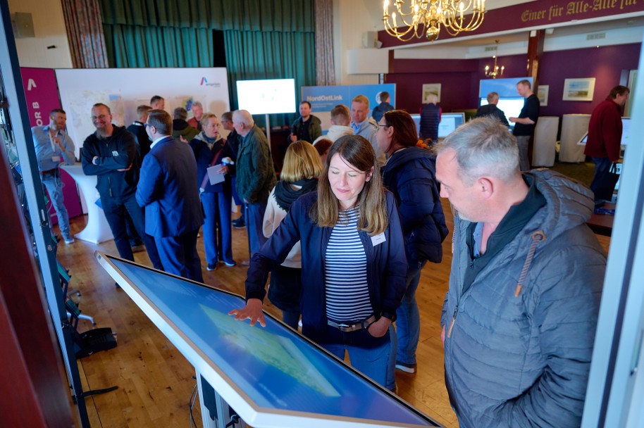 (c) Tennet/Timo Wilke
Im Vordergrund des Bildes stehen eine Frau und ein Mann an einem schräg gestellten Bildschirm, den die Frau mit der rechten Hand berührt. Sie scheint etwas zu zeigen. Im Hintergrund befinden sich jede Menge Menschen, die sich umsehen oder sich unterhalten.