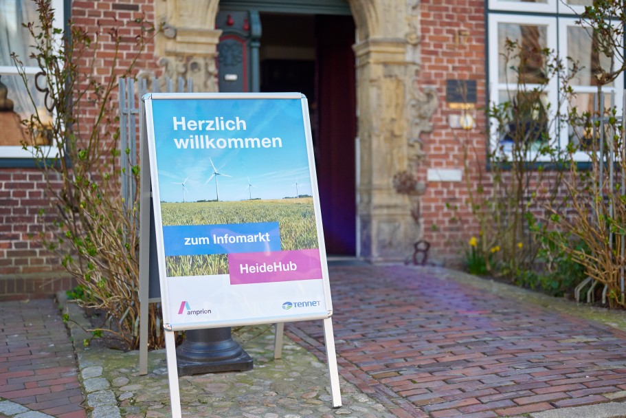 (c) Tennet/Timo Wilke
Ein aufklappbares Standschild mit der Aufschrift "Herzlich willkommen zum Infomarkt HeideHub" vor einer Eingangstür eines Backsteingebäudes