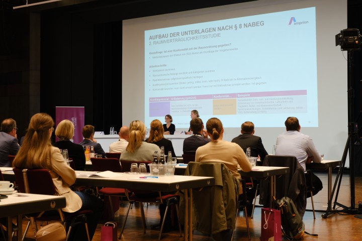 Vortragssituation: Auf einer Leinwand ist eine Präsentation mit Informationen zu sehen, vor der Leinwand stehem zwei Personen an Brückentischen. Noch weiter im Vordergrund sind einige Tischreihen zu sehen, an denen Menschen sitzen, die Richtung Leinwand blicken.