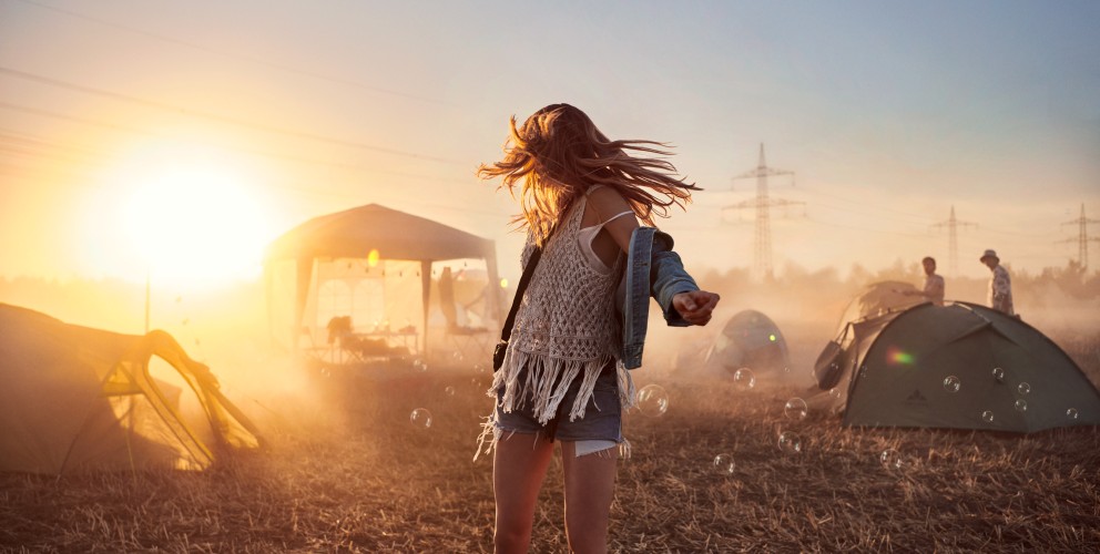 Das Bild zeigt eine junge Frau, die im Sonnenuntergang auf einer Wiese tanzt. Hinter ihr stehen einige Zelte und ein Pavillion. Im Hintergrund sind mehrere Strommaste zu sehen.