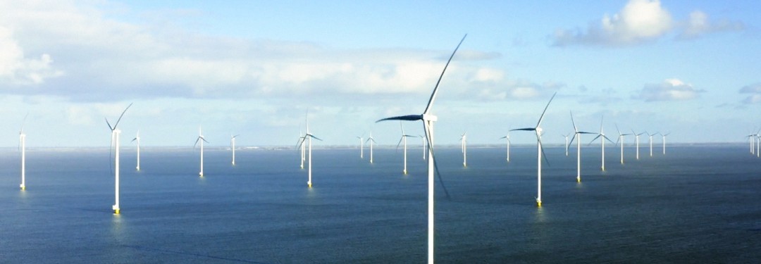 Mehrere Reihen mit Windrädern eines Offshore-Windparks. DAs Meer ist ruhig und dunkelblau, am Himmel einige Woken, am Horizont ist Land zu erkennen.