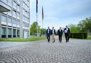 Vier Menschen laufen nebeneinander Richtung Kamera, einer hält einen Ordner im Arm.