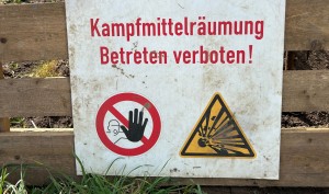Ein an einem Bretterzaun befestigtes Schild mit der roten Aufschrift "Kampfmittelräumung Betreten verboten" und zwei Warnhinweisen