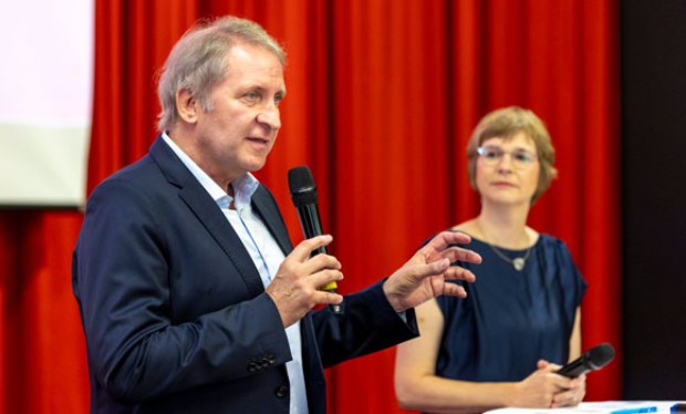 Ein Mann mit grauen Haaren und einem dunkelblauen Anzug spricht in ein Mikrofon und gestikuliert mit der anderen Hand. Hinter ihm steht eine Frau mit kurzem, blondem Haar und Brille, die ein Mikrofon hält und aufmerksam zuhört. Sie trägt ein dunkelblaues Kleid. Im Hintergrund hängt ein roter Vorhang, was auf eine Bühne oder einen Vortragssaal hindeutet.