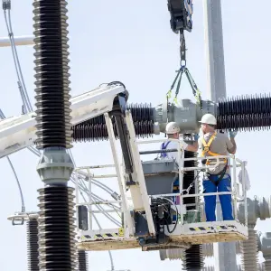 Zwei Arbeiter mit Sicherheitsausrüstung montieren ein Hochspannungs-Bauteil in einer Umspannanlage mithilfe eines Krans, stehend auf einer Arbeitsbühne.