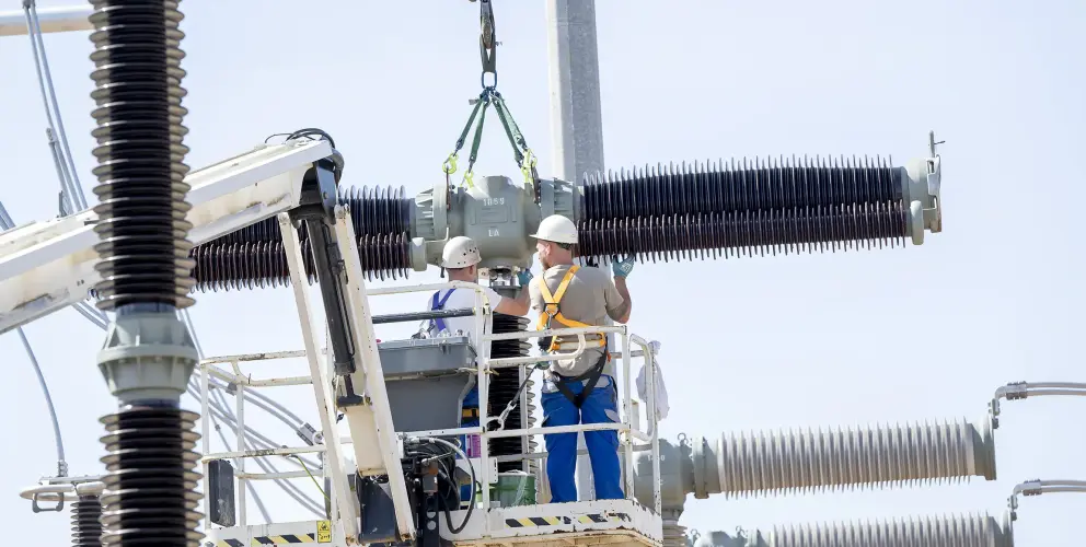 Zwei Arbeiter mit Sicherheitsausrüstung montieren ein Hochspannungs-Bauteil in einer Umspannanlage mithilfe eines Krans, stehend auf einer Arbeitsbühne.