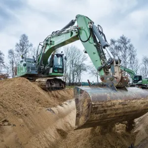Ein großer grüner Bagger bewegt Sand auf einer Baustelle in ländlicher Umgebung, im Hintergrund weitere Baumaschinen und kahle Bäume.