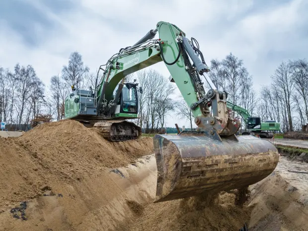 Ein großer grüner Bagger bewegt Sand auf einer Baustelle in ländlicher Umgebung, im Hintergrund weitere Baumaschinen und kahle Bäume.
